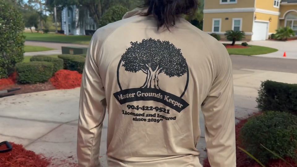 A Master Grounds Keeper crew member seen from behind, wearing a company shirt printed with the logo, phone number and “Licensed and Insured since 2019”.